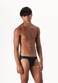 Maillots de bain noirs avec une texture lisse, dotés d'une large taille élastique. Le design comprend des côtés découpés pour une couverture minimale.