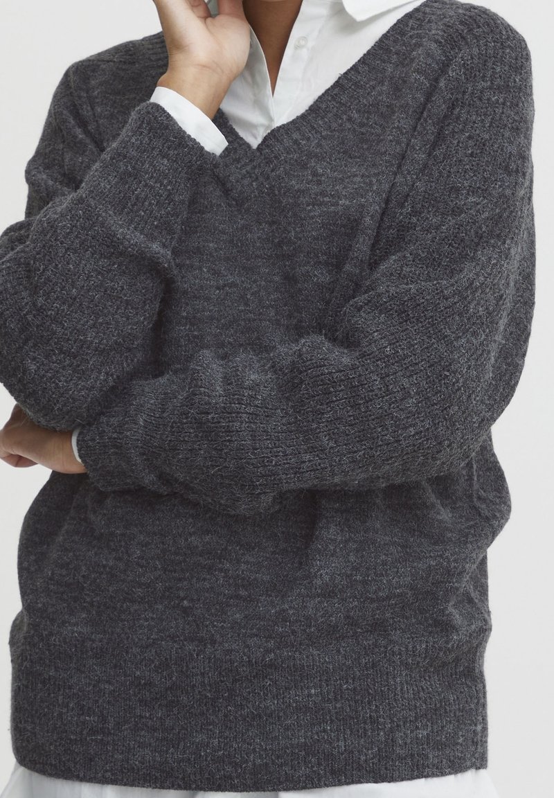 Personne portant un pull en maille gris foncé par-dessus une chemise blanche à col, les bras croisés, une main touchant le visage.