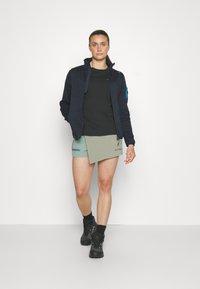 Sort zip-up jakke med blå detaljer, sort t-shirt og lys grøn skorts. Sorte vandrestøvler med hvide detaljer fuldender outfittet.