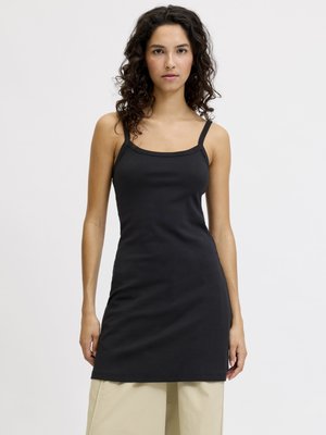 Frau mit lockigem dunklem Haar, die ein eng anliegendes schwarzes ärmelloses Kleid über beigen weiten Hosen vor einem schlichten Hintergrund trägt.