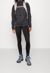 Czarna polarowa kurtka zapinana na zamek z wysokim kołnierzem, noszona na czarnych legginsach. Szare buty trekkingowe z niebieskimi akcentami i fakturowane skarpetki. Mały plecak przymocowany.