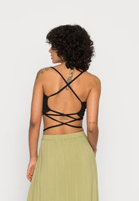 Bralette preto com alças cruzadas intrincadas nas costas; modelo veste uma saia verde fluida. Textura suave e material leve.