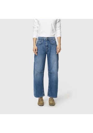 Kvinde iført højttaljede, vidde, blå jeans, hvid langærmet top og brune mokasinsko mod en ensfarvet grå baggrund.