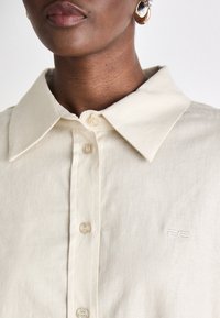 Beige button-up shirt with a pointed collar, textured fabric, and subtle embroidered logo on the left side. Smooth, clean design.