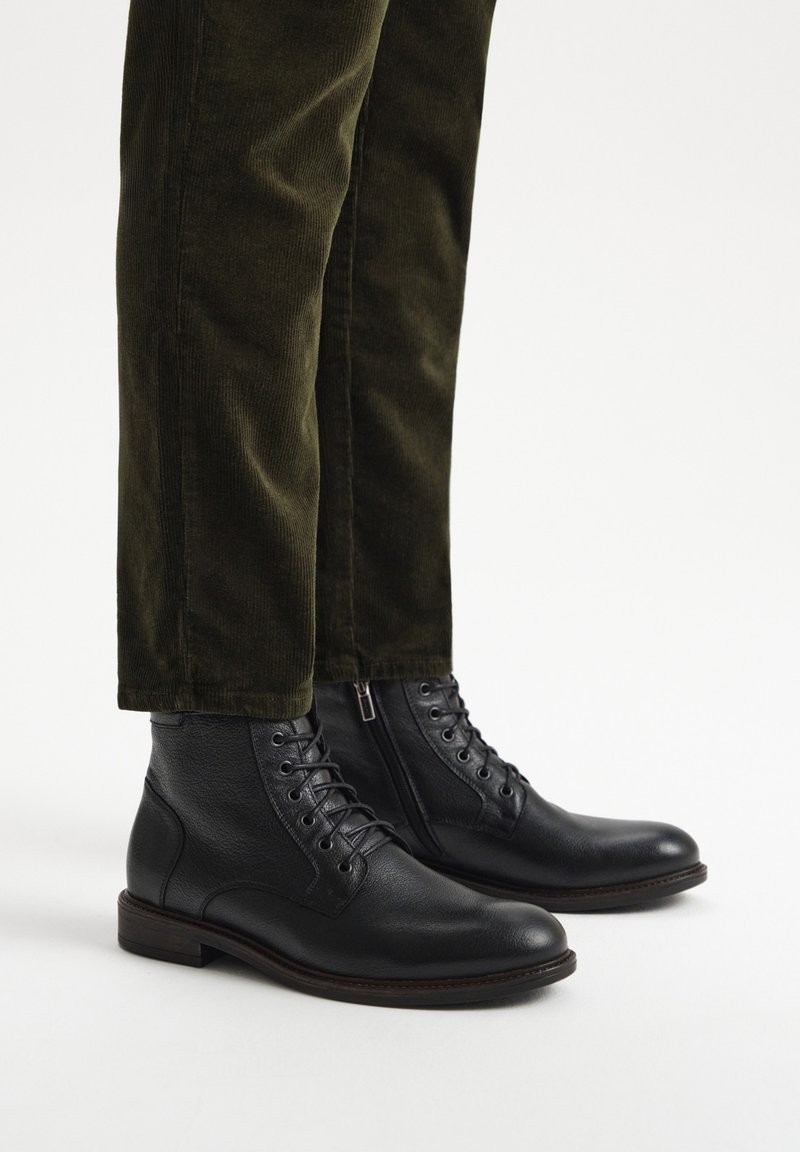 Botas de cuero negro hasta el tobillo con cordones, punta redonda y suela marrón; combinadas con pantalones de pana verde oliva que tienen un ligero brillo.