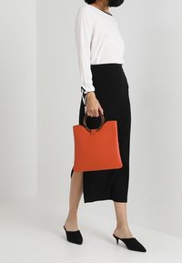 Rote Leder-Umhängetasche mit einem eleganten Design, ausgestattet mit einem Holzgriff und einer flachen, strukturierten Form, kombiniert mit einem weißen Oberteil und einem schwarzen Rock.