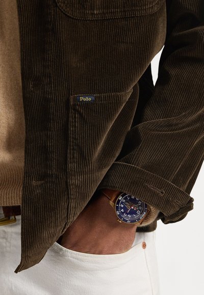 Καφέ ζακετάκι από κορντόροι με ετικέτα τσέπης "Polo", μπεζ πουλόβερ από κάτω και μπλε ρολόι με στρογγυλό καντράν και δερμάτινο λουρί. Ορατό άσπρο παντελόνι.