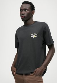 Melna kokvilnas T-kreklu ar aploksni, kurā ir krāsains "Rip Curl" logo uz krūšu. Modelis stāv ar rokām kabatās, ģērbies brūnās biksēs.