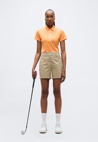 Femme debout tenant un club de golf, portant un polo orange, un short beige, des chaussettes blanches et des chaussures de golf blanches sur un fond uni.