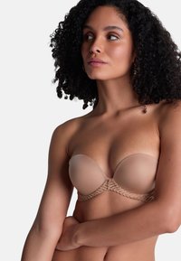 Femme aux cheveux bouclés portant un soutien-gorge nude sans bretelles avec des détails en dentelle, regardant sur le côté devant un fond blanc uni.
