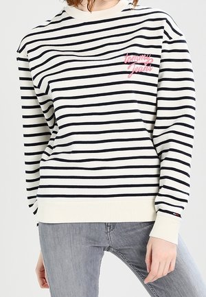 Osoba nosząca białą i czarną koszulę w poziome paski z długim rękawem i różowym tekstem "tommy jeans" na piersi oraz jasnoszare dżinsy.