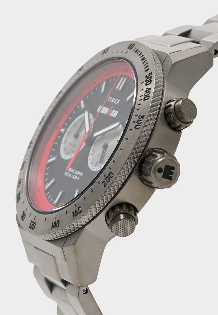 Primer plano de un reloj cronógrafo Timex Ironman plateado con esfera negra y roja, dos subesferas y botones laterales texturizados en una correa metálica.