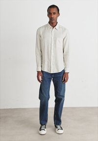 Camisa de lino beige claro de manga larga con un bolsillo en el pecho; vaqueros de mezclilla azules; zapatillas negras y blancas; de pie contra un fondo neutral.