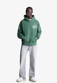 Zeleni bombažni hoodie s žepom, grafičnim besedilom "Srčka se ne zmotita," v paru z svetlo sivo jeans hlačami in belimi supergami.