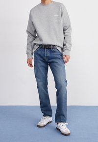 Sudadera gris de algodón con cuello redondo, vaqueros azules de denim, cinturón negro con ojales y zapatillas blancas con detalles marrones.