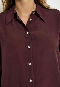 Blusa bordeaux con chiusura frontale a bottoni, texture morbida, colletto classico e bottoni bianchi. Il tessuto appare leggero e leggermente trasparente.