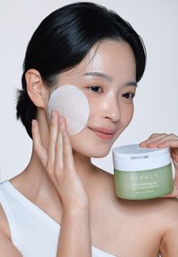 Vihreä purkki, jossa lukee "NEEDLY Cicachid Chilling Pad", pidetään henkilön vieressä, joka käyttää valkoista pyöreää vanulappua poskelleen. Valkoinen tausta.