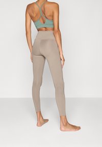 Světl zelená sportovní podprsenka s výřezem na zádech spárovaná s nahými, vysokými legínami. Tkanina vypadá hladce a elasticky.