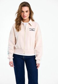 Žena s vlnitými vlasmi oblečená v svetlo béžovej mikine na zips s nápisom "MADE OF ECHOS AND SLOW HORIZONS" a tmavomodrých džínsoch stojí pred bielym pozadím.