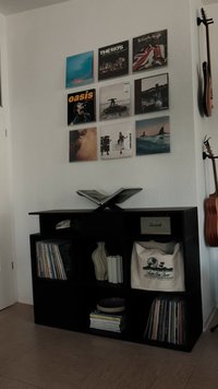 Svart trebokhylle med en åpen bok, vinylplater, en dekorativ vase og en beige veske. Veggen har innrammede albumcover over hyllen.