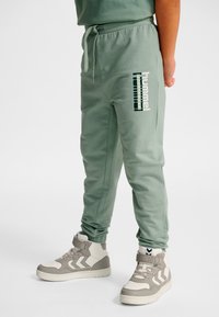 Kind draagt groene joggingbroek met "hummel sportswear" logo en wit-grijze hoge sneakers, staand op een witte achtergrond.