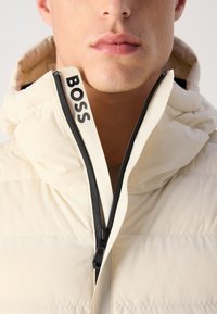 Close-up van een man in een beige gewatteerde jas met een zwarte rits en een gedeeltelijk dichtgeritste kraag met het logo "BOSS".