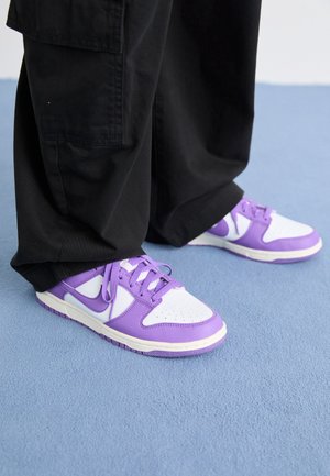 Osoba ubrana w czarne spodnie o szerokich nogawkach i białe trampki z fioletowymi akcentami, stojąca na niebieskim, wyściełanym dywanie.