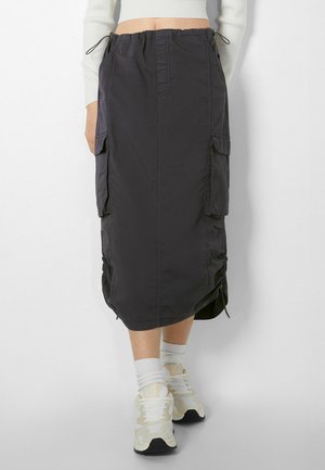 Zwart cargo rokje van middellange lengte met zijzakken en elastische tailleband, gedragen met witte sokken en beige sneakers, tegen een effen achtergrond.