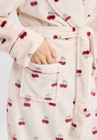 Albornoz de forro polar rosa con un patrón de cerezas, que incluye un cinturón en la cintura y un bolsillo parche en la parte delantera. Textura suave con detalles en rojo que contrastan.