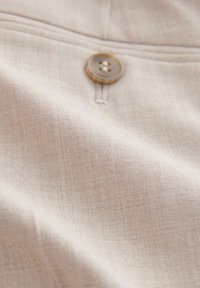 Tessuto beige chiaro con una trama sottile, caratterizzato da un bottone marrone cucito al centro. Primo piano di un materiale morbido e liscio.