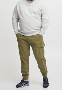 Grijze sweatshirt van een gestructureerde stof, olijfgroene cargobroek met zijzakken, en blauwe sportschoenen met witte accenten.