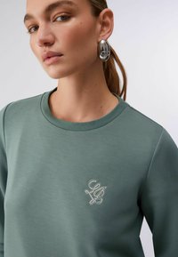 Sweat-shirt vert à encolure ronde, avec un logo orné de strass sur la poitrine. Le tissu a une texture lisse. Boucles d'oreilles argentées visibles.