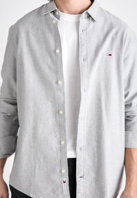 Uomo che indossa una camicia grigia chiaro con bottoni sopra una maglietta bianca, maniche arrotolate, con un piccolo logo rosso, bianco e blu sul petto e sull'orlo.