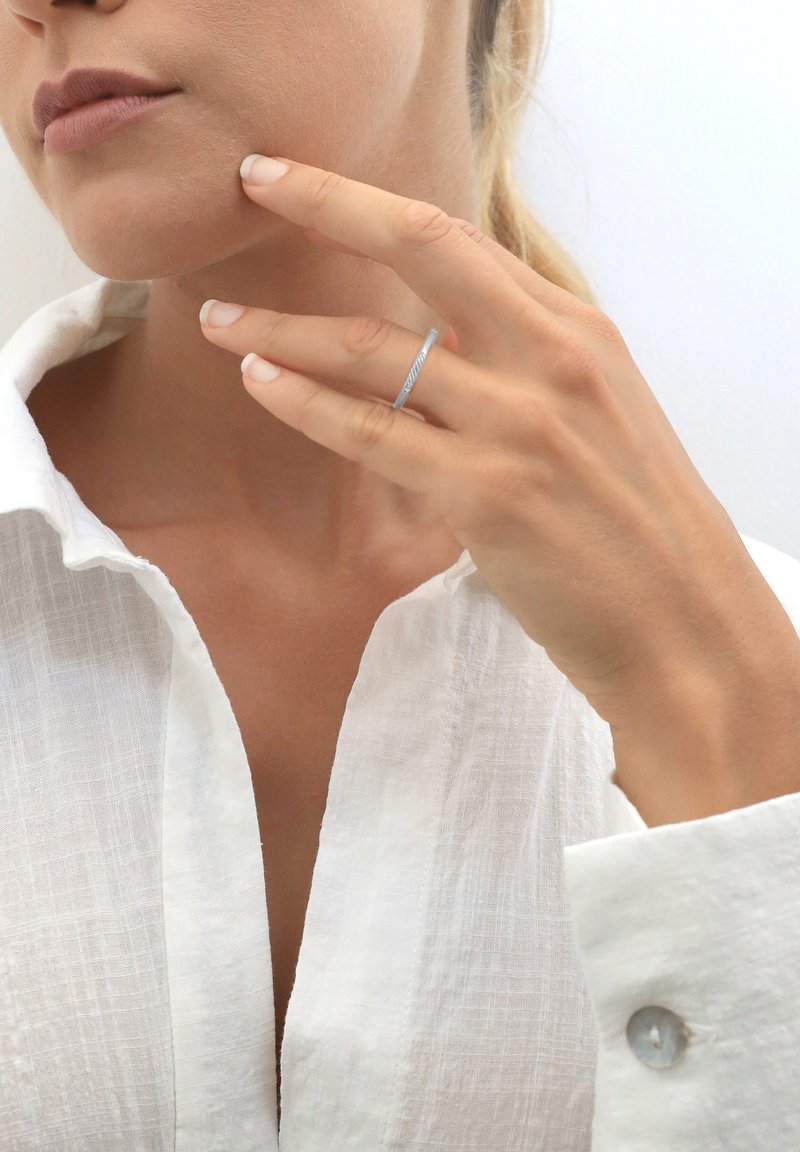 Sottile anello d'argento con un motivo diagonale testurizzato, indossato sull'anulare, su uno sfondo di una camicia di lino bianca e tono della pelle.