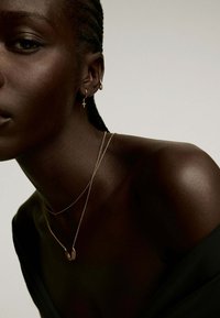 Primer plano del cuello de un modelo que exhibe collares de oro en capas y pendientes de aro. La piel es de un tono marrón oscuro, con trenzas texturizadas visibles. Ropa negra.