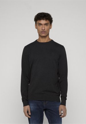 Vīrietis ar cirtainiem matiem, valkājot melnu džemperi ar apaļu kaklu un zilas džinsas, stāv pret vienkāršu gaiši pelēku fonu.