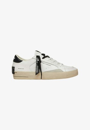 Sneaker bianco con accenti neri, caratterizzato da una tomaia in pelle liscia, lati testurizzati, lacci piatti e una suola in gomma beige spessa. Etichetta del marchio visibile.