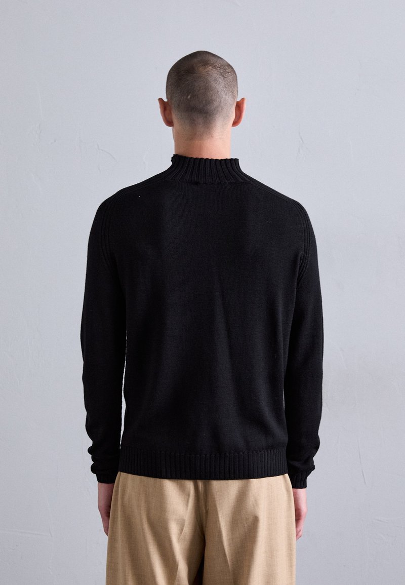 Mann mit kurzem Haar, der wegschaut, trägt einen schwarzen Strickpullover und beige Hosen vor einem schlichten grauen Wandhintergrund.