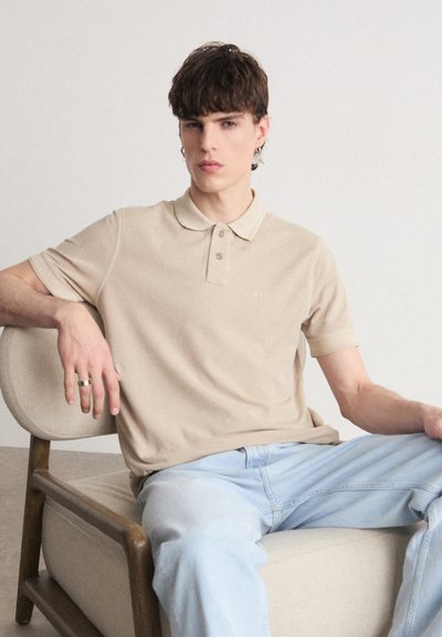 Polo en coton beige avec un col classique, patte de boutonnage à trois boutons et logo brodé. Jean en denim bleu clair, assis sur une chaise en bois.