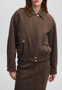 Bruin suède cropped jack met een kraag, drukknopen aan de voorkant, twee borstzakken en een elastische taille met geribbeld patroon.