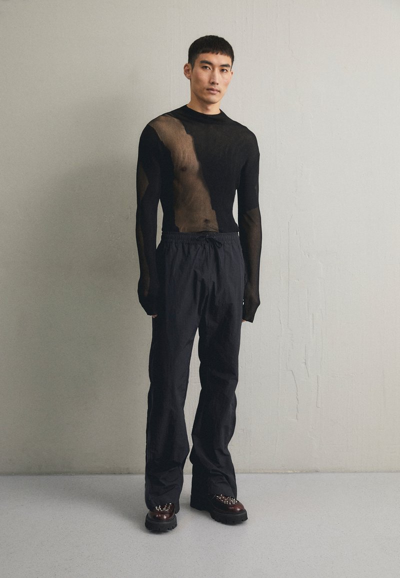 Top lungo a maniche nere trasparenti con una finitura testurizzata, abbinato a pantaloni neri larghi e stivali chunky con decorazioni metalliche.