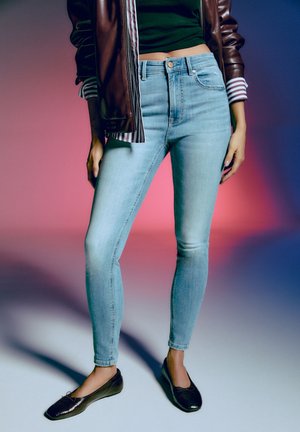 Woman wearing light blue skinny jeans, black ballet flats, a black cropped top, and a brown leather jacket with striped cuffs.