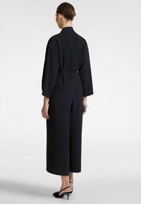 Mustapuku, jossa on pitkät hihat, vyötärönauha, leveät lahkeet ja korkea kaula-aukko. Sujuva tekstuuri ja minimalistinen muotoilu hienovaraisilla yksityiskohdilla.