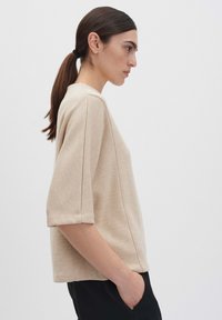Beiger Kurzarmpullover mit entspannter Passform aus strukturiertem Stoff. Verfügt über Seitennähte und einen Rundhalsausschnitt. Modell posiert seitlich.