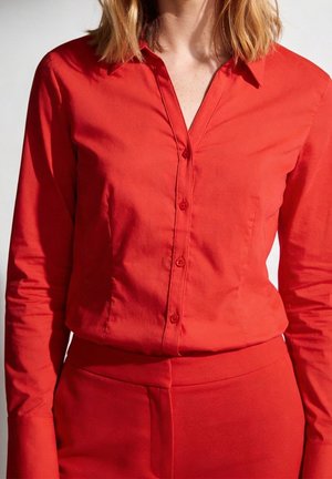 Femme portant une chemise rouge ajustée à manches longues boutonnée, rentrée dans un pantalon rouge taille haute, sur un fond uni.