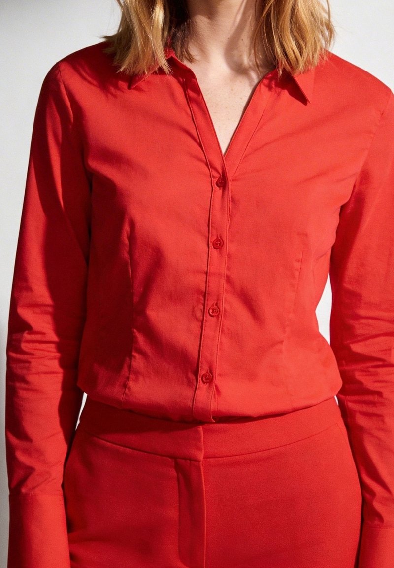 Femme portant une chemise rouge ajustée à manches longues boutonnée, rentrée dans un pantalon rouge taille haute, sur un fond uni.