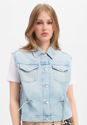 Junge Frau mit hellbraunen Haaren trägt ärmellose hellblaue Jeansjacke über weißem Hemd vor einfachem Hintergrund.