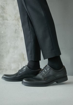 Persona con zapatos negros de vestir con cordones, calcetines negros y pantalones negros a medida, de pie sobre un suelo gris claro frente a una pared texturizada.