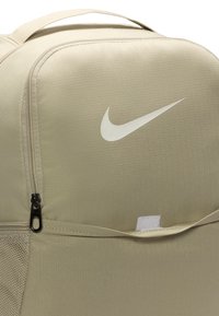 Mochila Nike beige con una textura de tela suave, que presenta un bolsillo con cremallera y un logotipo blanco prominente. Correa ajustable y detalles de malla visibles.