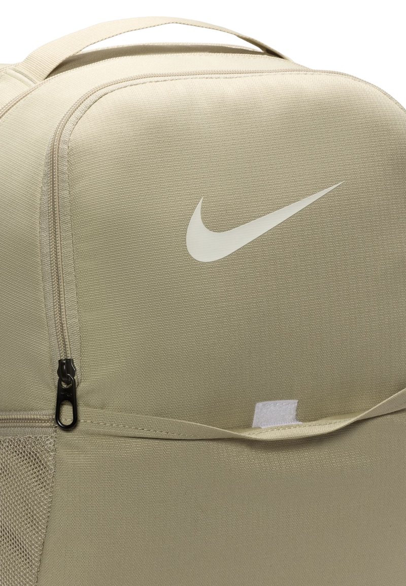 Beige Nike ryggsäck med en slät tygtextur, utrustad med en dragkedjeficka och en framträdande vit logotyp. Justerbar rem och meshdetaljer synliga.